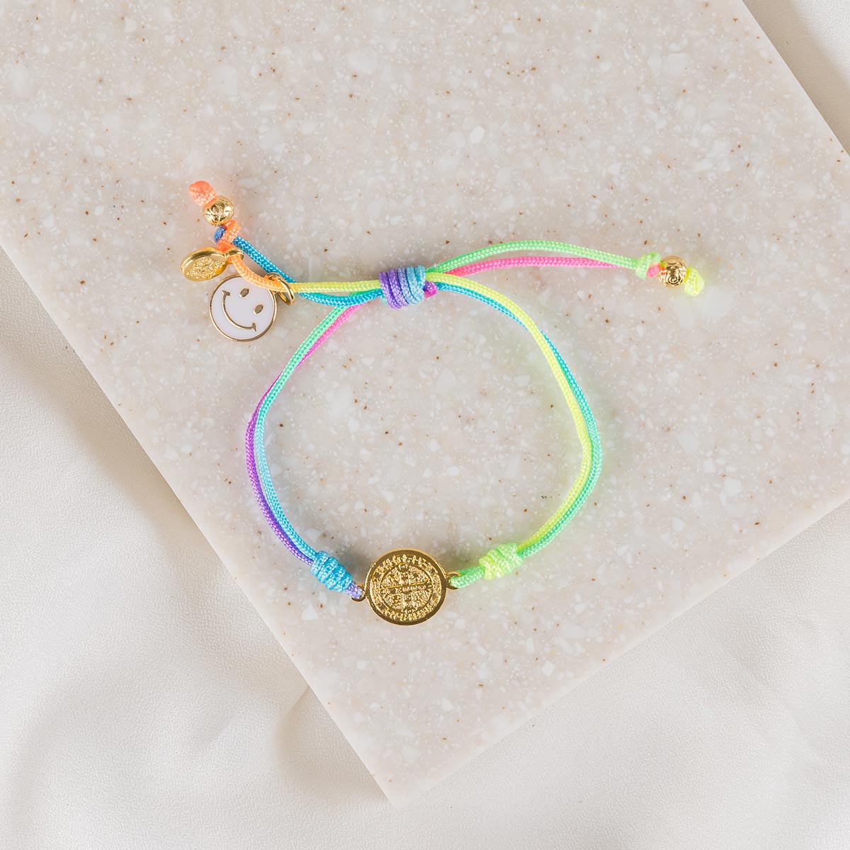 A colorful woven bracelet with a gold-tone Benedictine Medal and a 'blessed' tag, featuring a happy face charm and rainbow threading, presented on a neutral background.