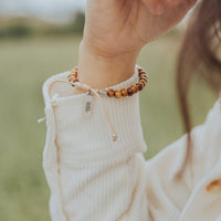 A close-up of a person's wrist wearing a beaded bracelet with a cream cord and a small tag that reads 'FEEL'. The person is wearing a cream ribbed knit sweater, and the background is a soft-focus outdoor grassy field.