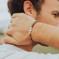 A close-up lifestyle shot of a man wearing a beaded bracelet on his wrist. The bracelet features brown wooden beads and a small silver cross accent, worn while the man holds his neck.