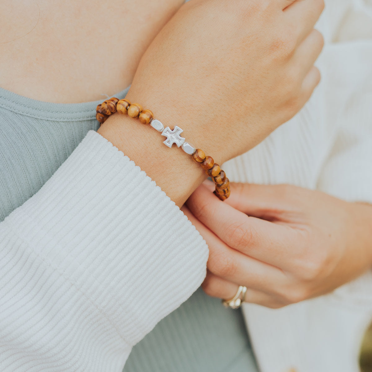 A close-up of a person's wrist wearing a beaded bracelet with brown wooden beads and a silver cross charm, paired with a white ribbed sweater.