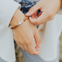 A close-up lifestyle shot of a person wearing a beaded bracelet with a silver puzzle piece and silver oval charm, while another person adjusts the bracelet.