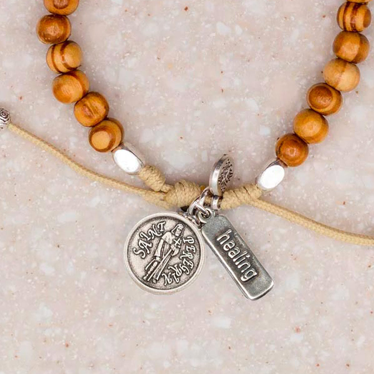 A close-up of a beaded cord bracelet with warm brown wooden beads, a tan cord, white spacer beads, and silver-tone charms including a round Saint Peregrine medal and a rectangular tag engraved with the word 'healing'.