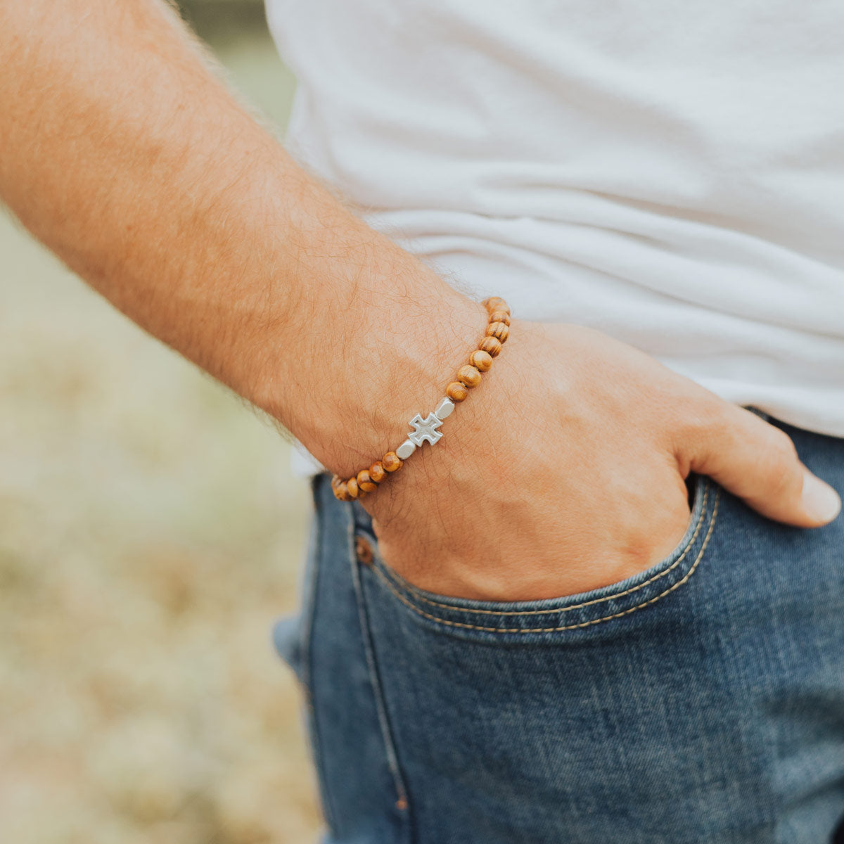 A close-up of a man's hand in his jeans pocket wearing a beaded bracelet with a silver cross charm.