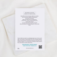 A printed prayer card titled 'Archangel Michael Prayer of Protection' with a short prayer and longer explanatory text, placed on a soft white fabric background. The card includes the brand name Wearable Blessings and a QR code.