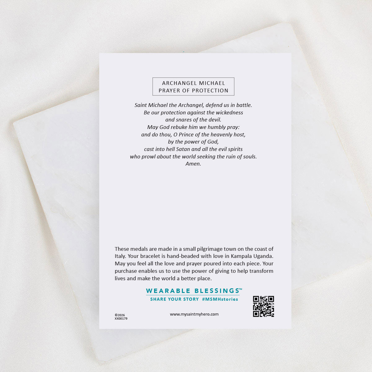 A printed prayer card titled 'Archangel Michael Prayer of Protection' with a short prayer and longer explanatory text, placed on a soft white fabric background. The card includes the brand name Wearable Blessings and a QR code.