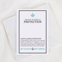 A white rectangular card with black and teal text reading 'SAINTS & ANGELS PROTECTION' and 'MY SAINT, MY HERO.' is photographed on a soft white fabric background.