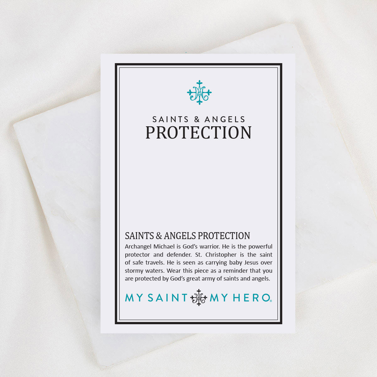 A white rectangular card with black and teal text reading 'SAINTS & ANGELS PROTECTION' and 'MY SAINT, MY HERO.' is photographed on a soft white fabric background.