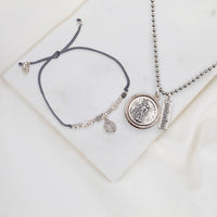 A jewelry set featuring a dark gray adjustable cord bracelet and a silver-tone necklace with a round medallion pendant and a small rectangular tag, displayed on a white marble tile.