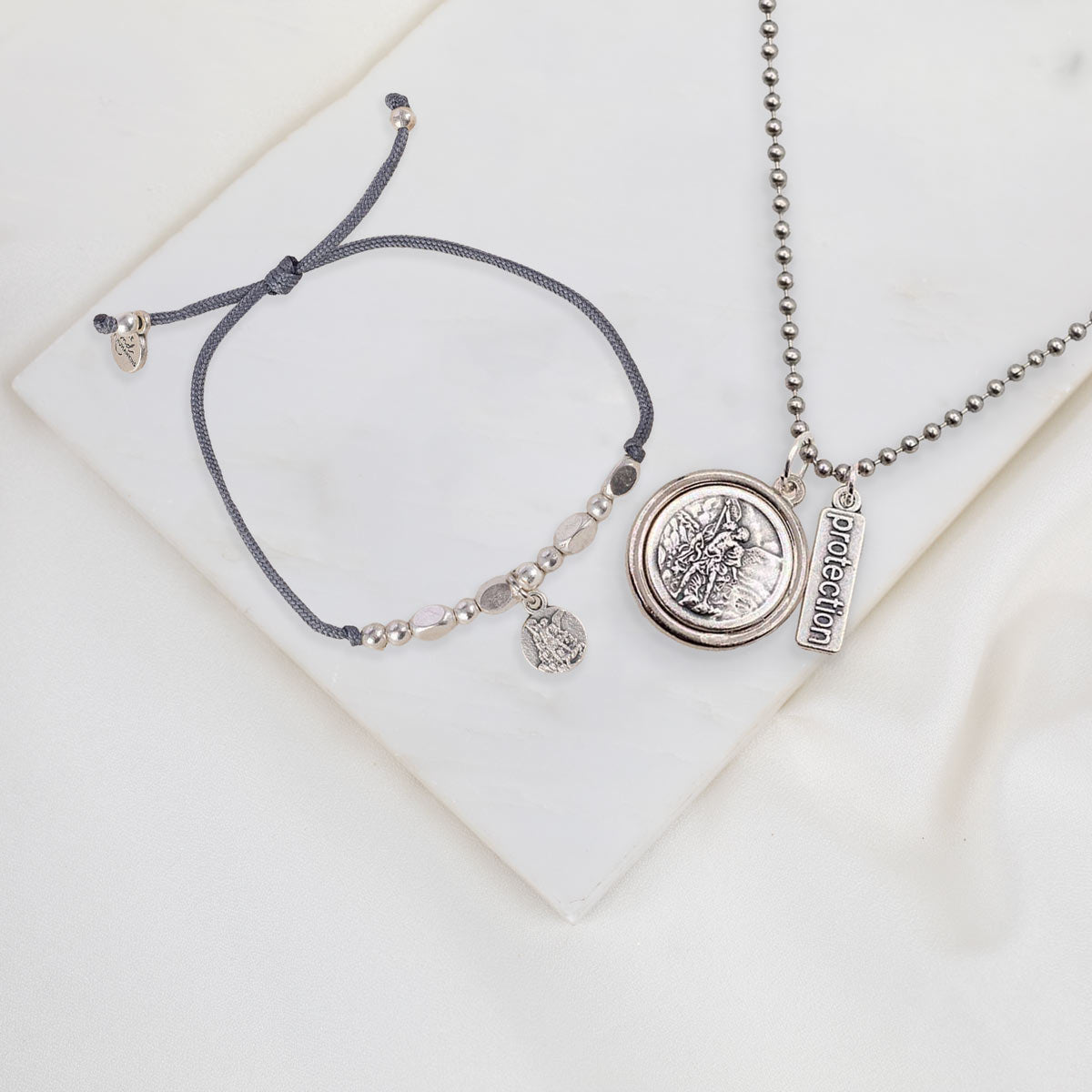 A jewelry set featuring a dark gray adjustable cord bracelet and a silver-tone necklace with a round medallion pendant and a small rectangular tag, displayed on a white marble tile.