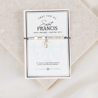 An adjustable slate grey bracelet with silver-tone metal beads and a St. Francis of Assisi medal, packaged on an inspirational card with the text 'PRAY FOR US' and the feast day October 4.