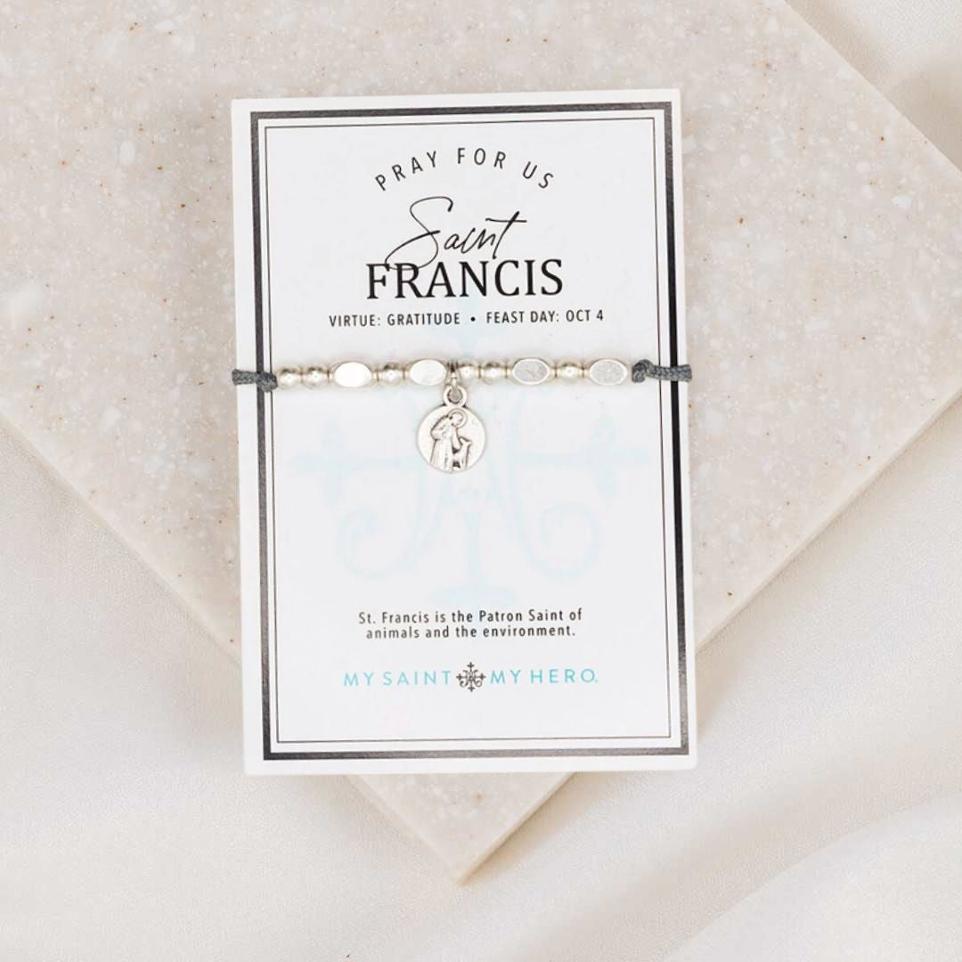 An adjustable slate grey bracelet with silver-tone metal beads and a St. Francis of Assisi medal, packaged on an inspirational card with the text 'PRAY FOR US' and the feast day October 4.
