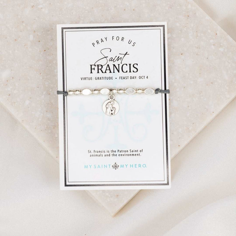 An adjustable slate grey bracelet with silver-tone metal beads and a St. Francis of Assisi medal, packaged on an inspirational card with the text 'PRAY FOR US' and the feast day October 4.