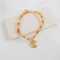 Beaded carnelian gemstone bracelet with a gold sand dollar charm on a white stone surface