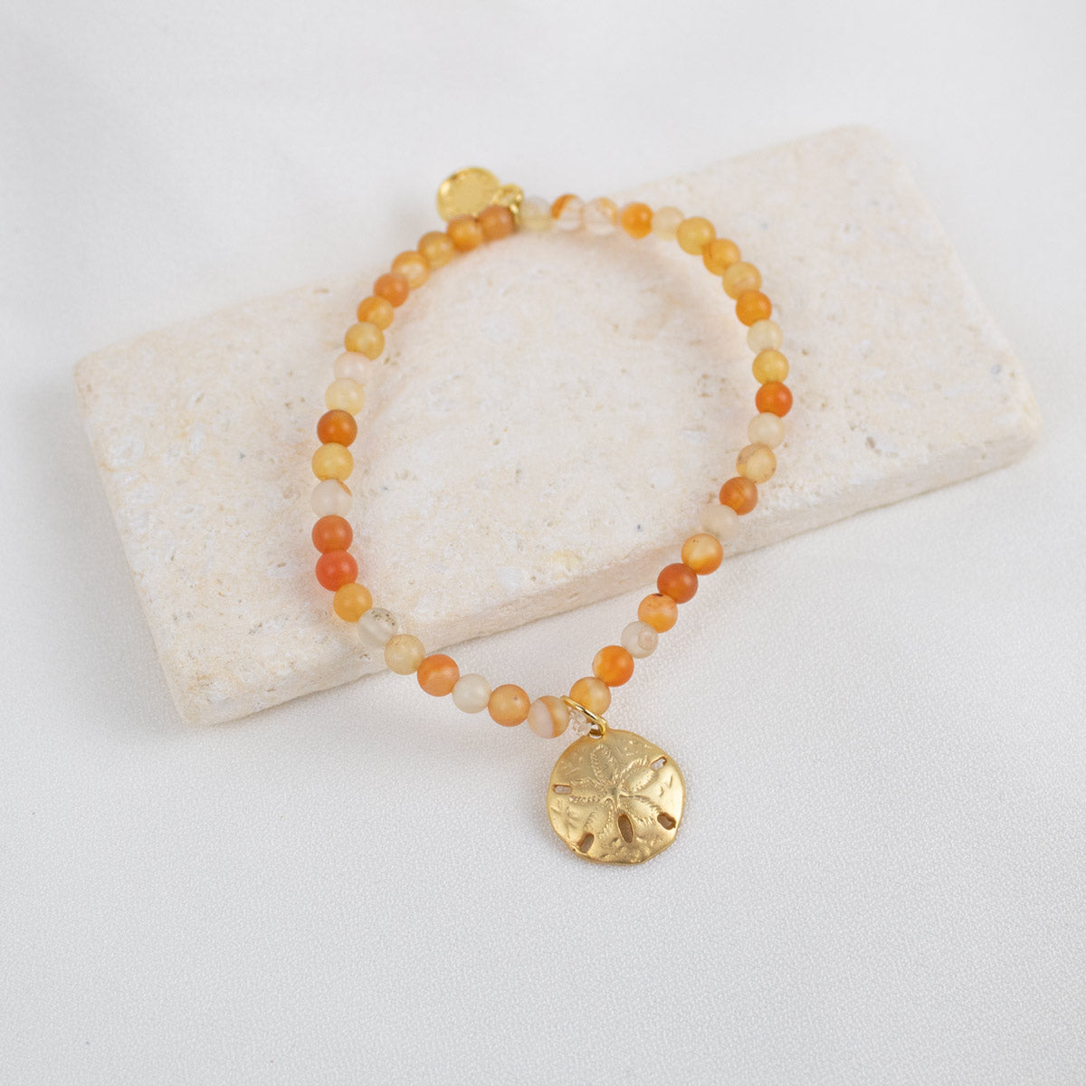 Beaded carnelian gemstone bracelet with a gold sand dollar charm on a white stone surface