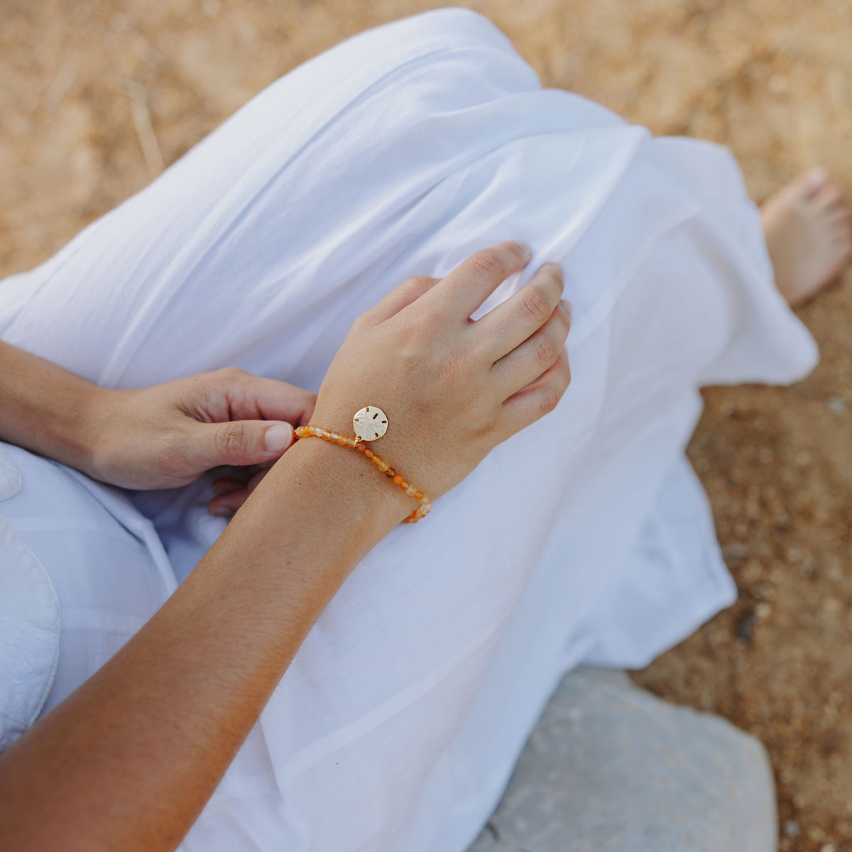 Person sitting on the beach wearing a white dress and orange bracelet with a sand dollar charm.