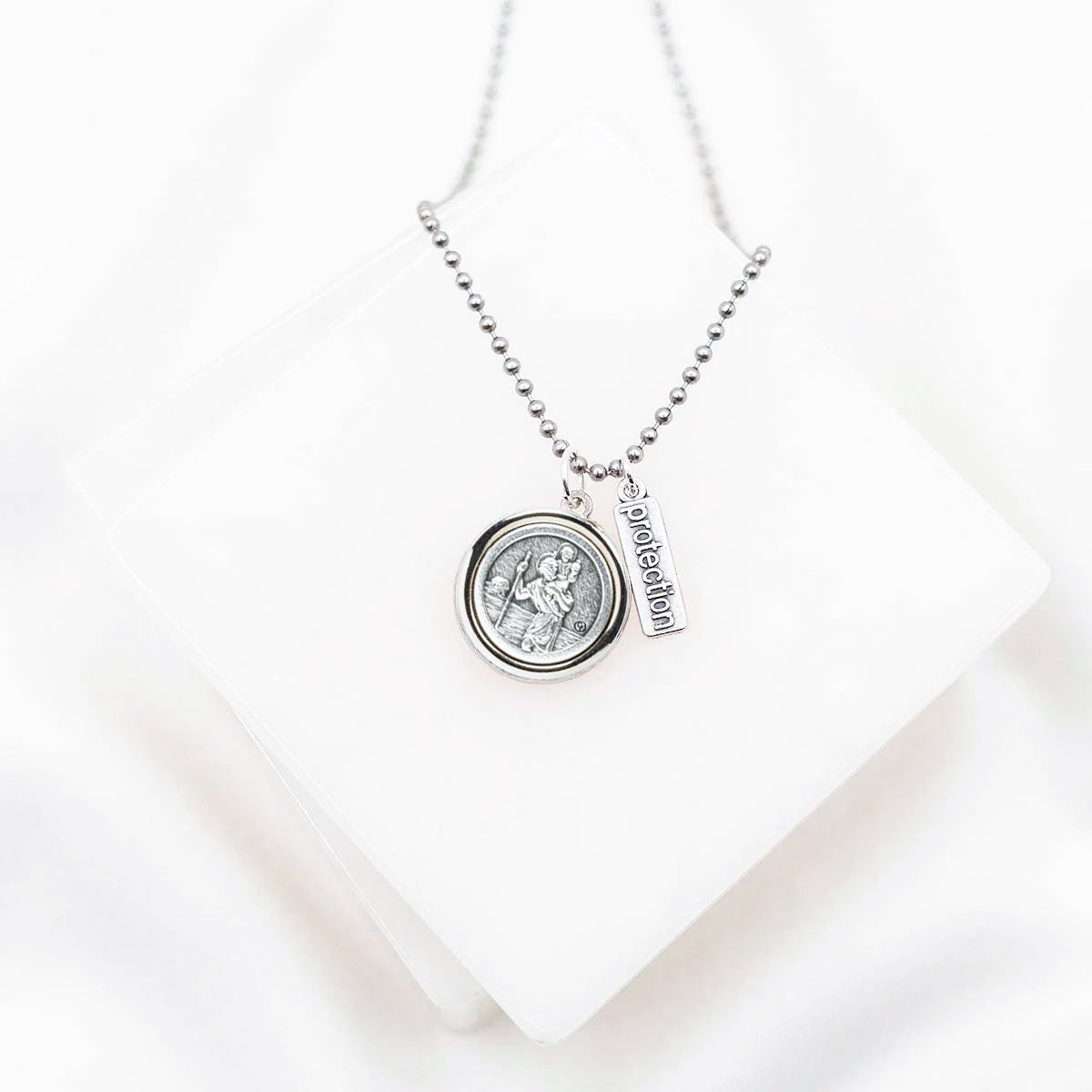 A silver necklace with a round St. Christopher/Archangel Michael medallion pendant and a vertical 'protection' charm, displayed on a white geometric block against a white background.