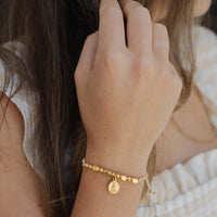 Close-up of a hand wearing a gold bracelet with a St. Bernadette Our Lady of Lourdes beaded Little Saints Bracelet against a blurred background.