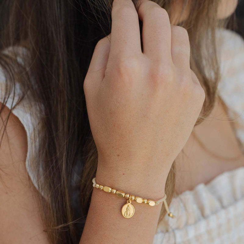 Close-up of a hand wearing a gold bracelet with a St. Bernadette Our Lady of Lourdes beaded Little Saints Bracelet against a blurred background.