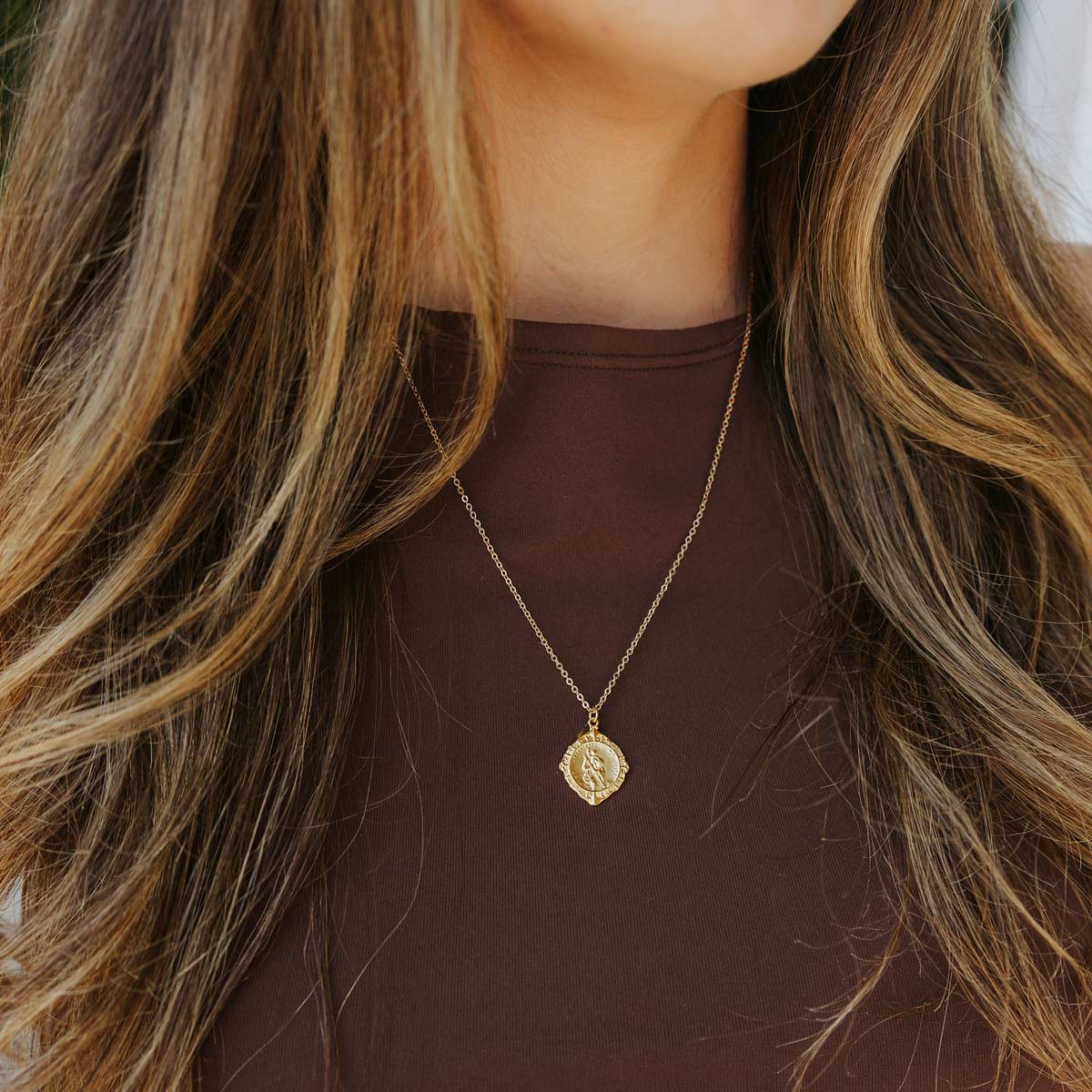 Gold necklace with a pendant worn by a person with long hair, against a neutral background.