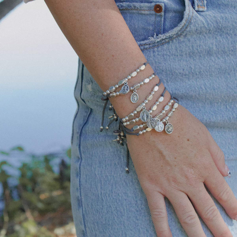 Woman's arm wearing a stack of little saints silver beaded and corded bracelets with saint medals