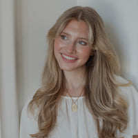 A smiling blonde woman with long wavy hair, wearing a white top and a gold pendant necklace, photographed against a soft off-white background.