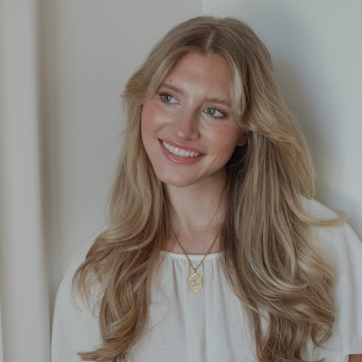 A smiling blonde woman with long wavy hair, wearing a white top and a gold pendant necklace, photographed against a soft off-white background.