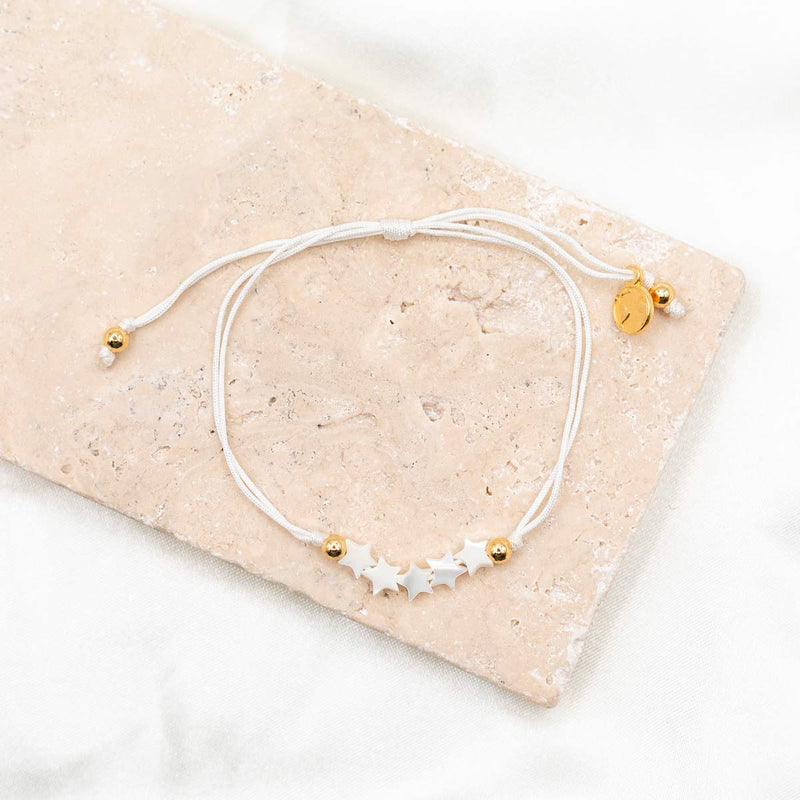 A white bracelet with gold-tone metal beads and star-shaped pendants, displayed on a beige surface.