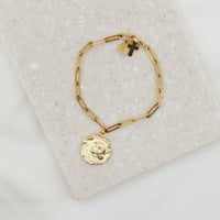 A gold-tone St. Joan of Arc bracelet with a paperclip link design and a medal charm, displayed on a beige surface.