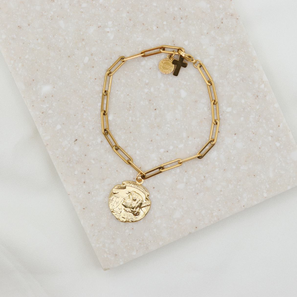 A gold-tone St. Joan of Arc bracelet with a paperclip link design and a medal charm, displayed on a beige surface.