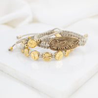 A bracelet stack of gold tone Angel Wing and floral medal bracelets on tan handwoven cording.