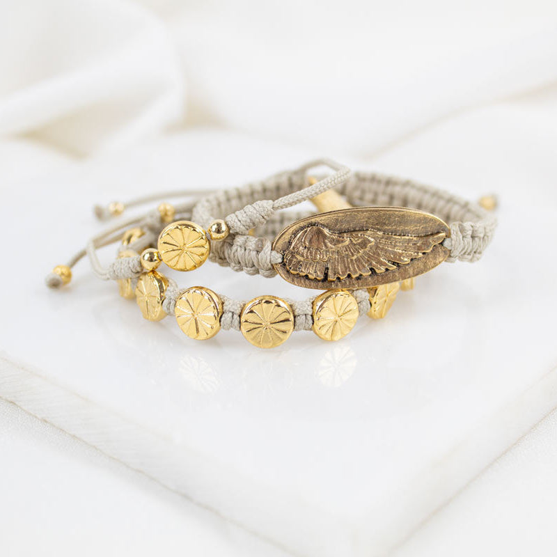 A bracelet stack of gold tone Angel Wing and floral medal bracelets on tan handwoven cording.