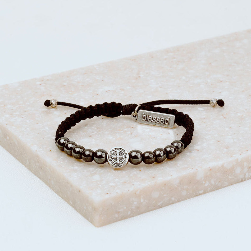10 hematite gemstones and a silver tone benedictine medal handwoven into a slip knot bracelet with a silver tone "blessed" charm.