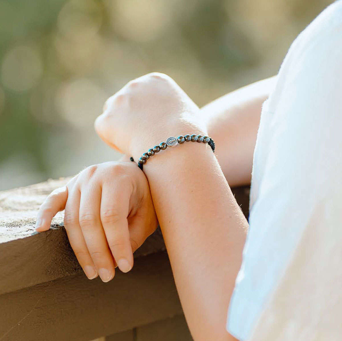 A close-up lifestyle shot of a person's hands and arms resting on a wooden railing, wearing a beaded bracelet with dark gray beads and a central silver-toned oval charm.