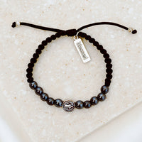 10 hematite gemstones and a silver tone benedictine medal handwoven into a slip knot bracelet with a silver tone "blessed" charm.