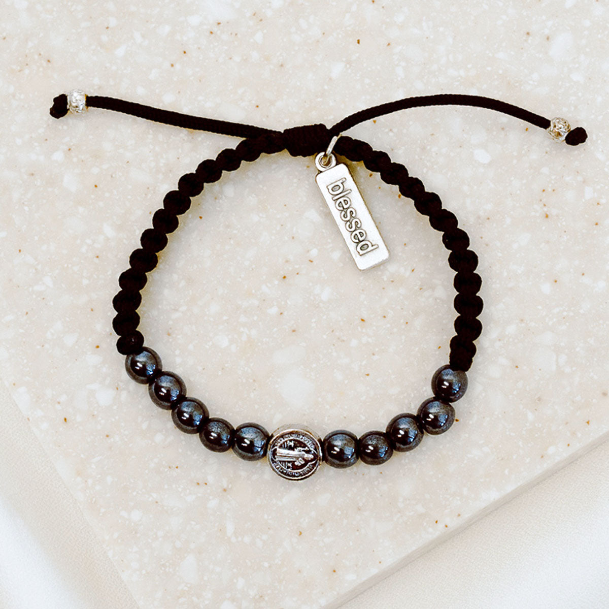 10 hematite gemstones and a silver tone benedictine medal handwoven into a slip knot bracelet with a silver tone "blessed" charm.