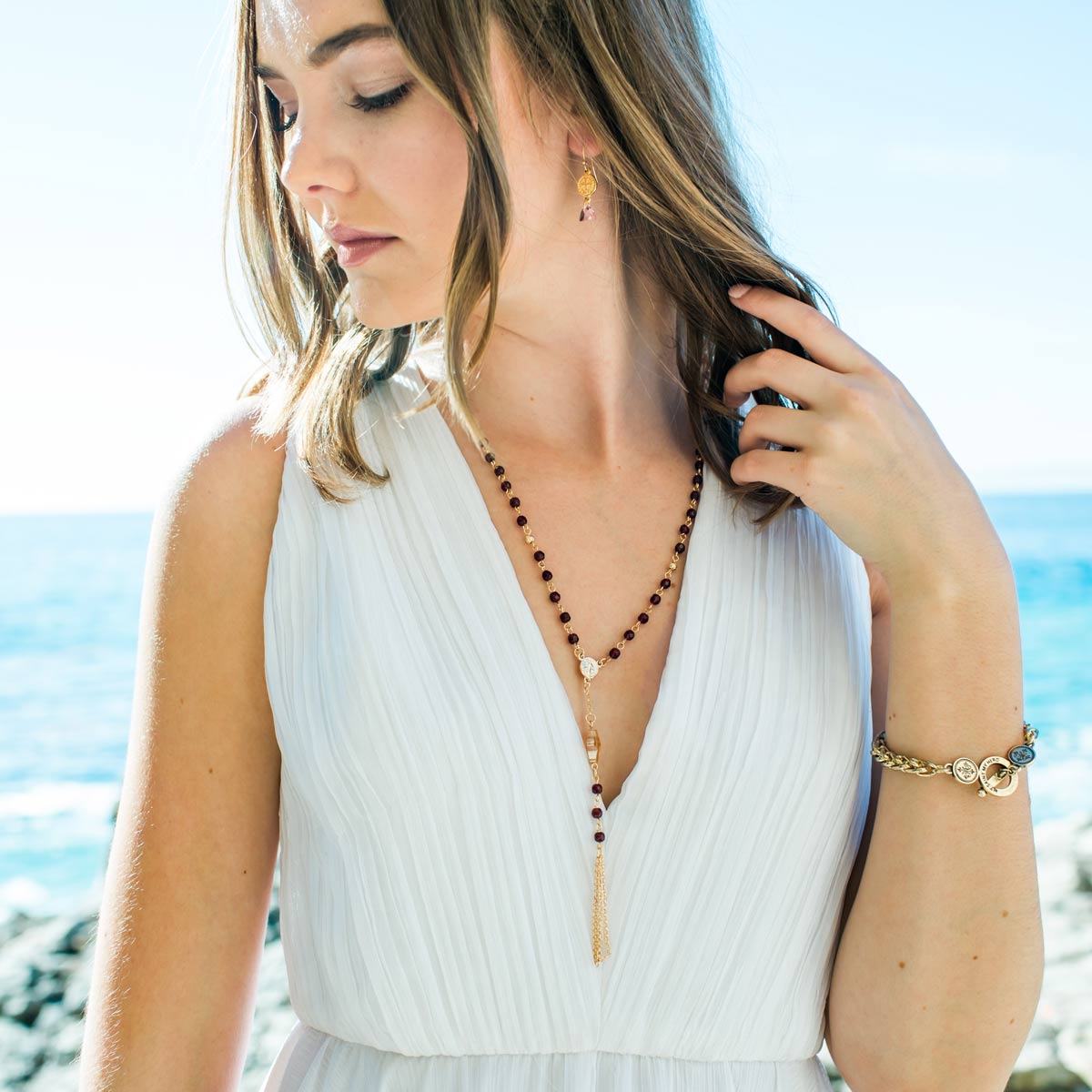 A woman wearing a white pleated sleeveless top and gold-toned jewelry, including a long beaded Rosary necklace with a tassel and a bracelet, standing outdoors near the ocean.