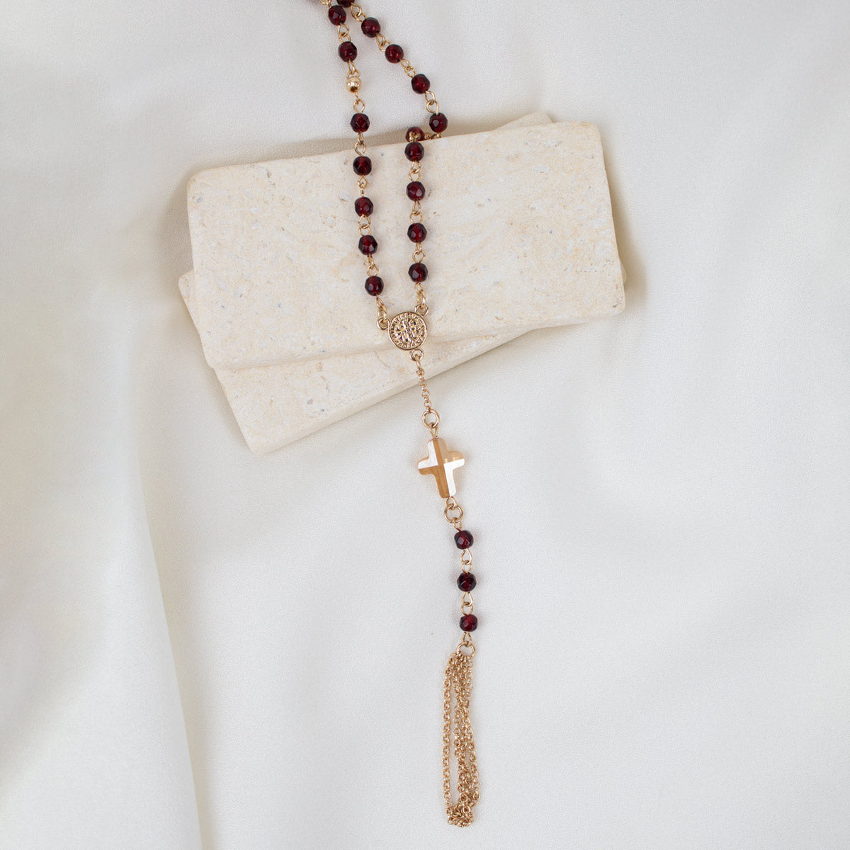 A rosary necklace with dark red beads and gold-toned metal links, cross, and tassel, draped over a folded cream-colored stone block on a white fabric background.