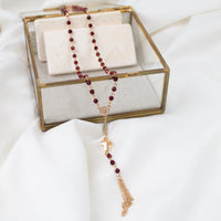 A rosary necklace with dark red beads and a gold-tone cross pendant, draped over a clear glass jewelry box with a white fabric background.