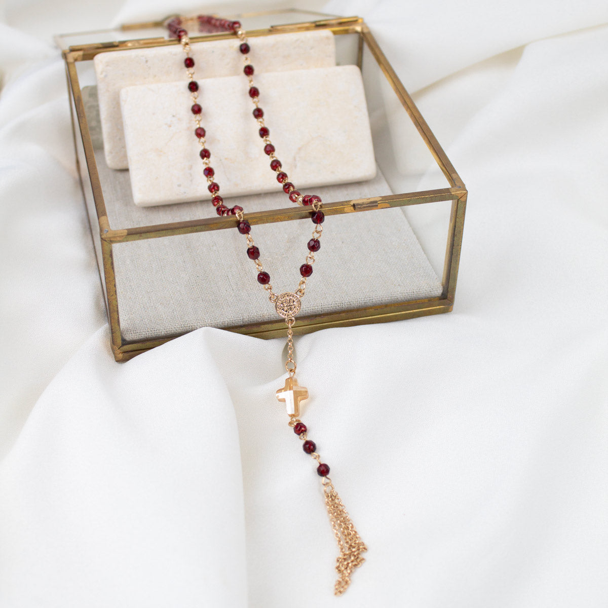 A rosary necklace with dark red beads and a gold-tone cross pendant, draped over a clear glass jewelry box with a white fabric background.