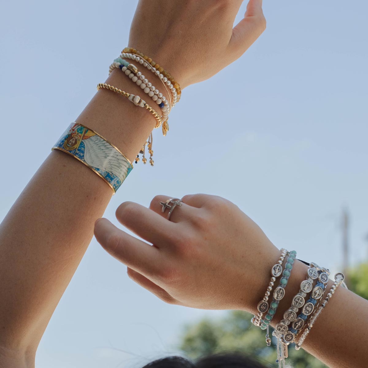 A close-up lifestyle shot of two human arms and hands adorned with multiple bracelets against a pale blue sky. The bracelets include a wide metallic cuff archangel Michael sacred image icon, several delicate beaded strands, silver-toned chains, and turquoise accents.