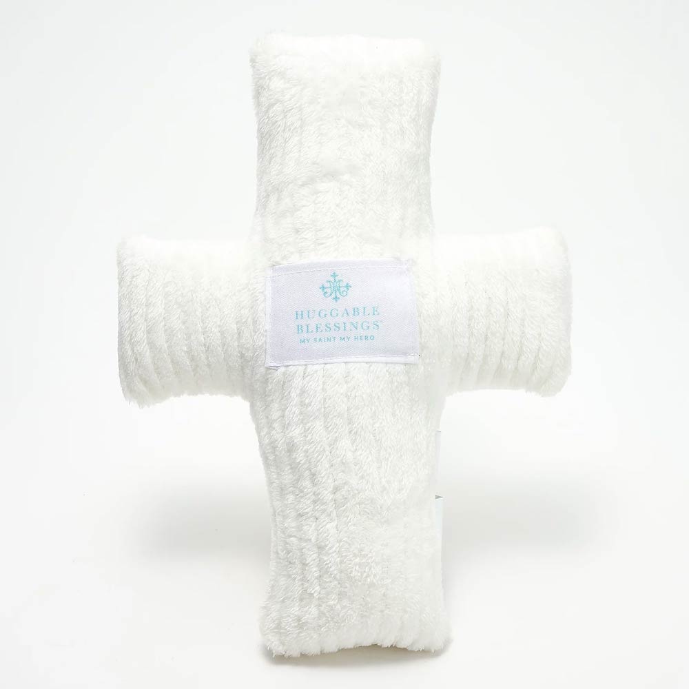 A plush, white weighted pillow in the shape of a cross with a label that reads 'Huggable Blessings'.