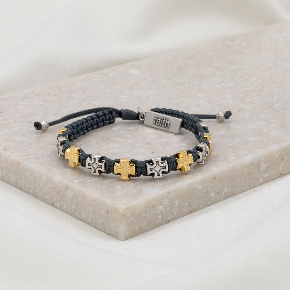 slate woven bracelet with alternating silver and gold tone medals and a silver charm tag at the slip knot closure with the word faith