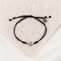 A black adjustable string bracelet featuring a silver-tone Benedictine Medal and a charm, displayed on a textured background.