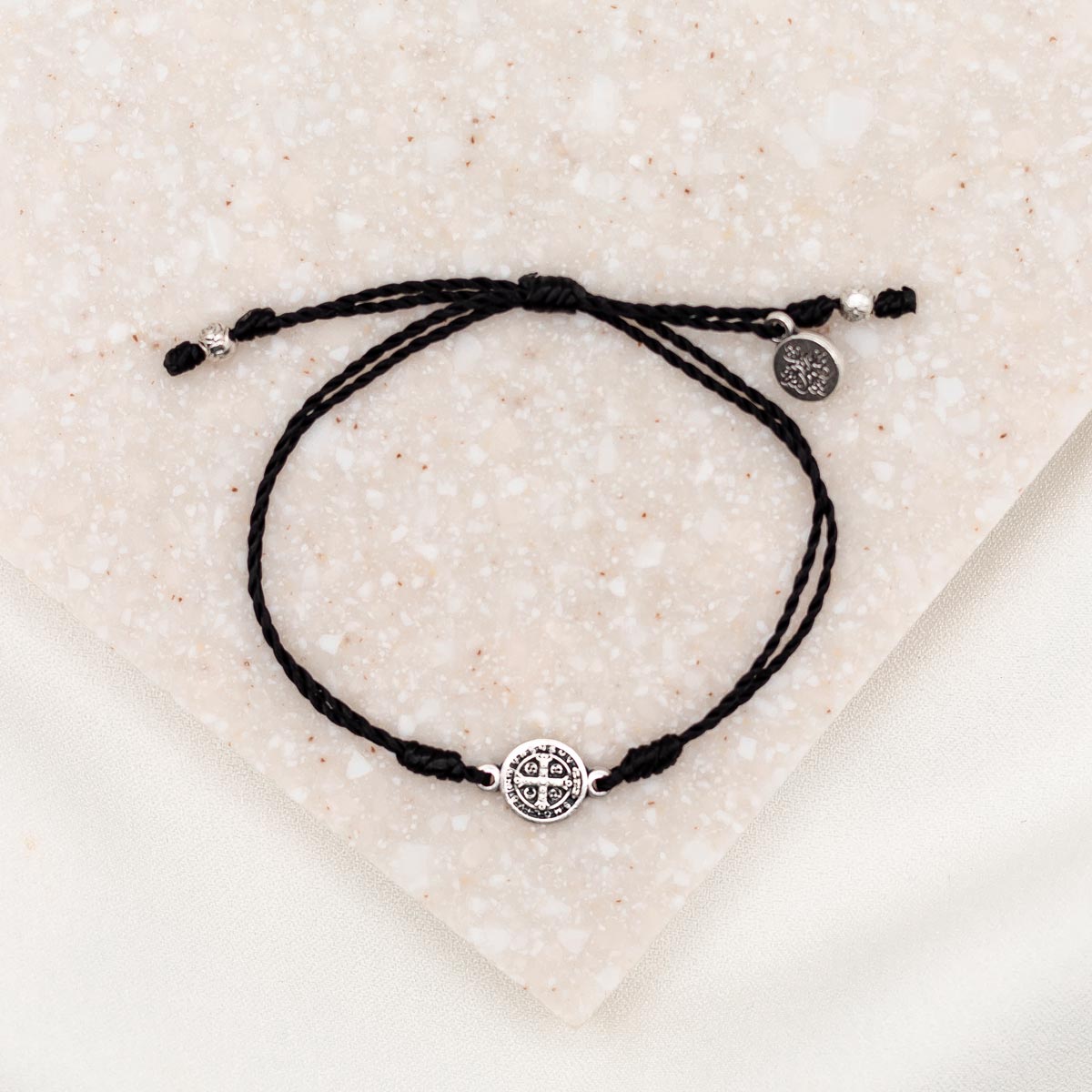 A black adjustable string bracelet featuring a silver-tone Benedictine Medal and a charm, displayed on a textured background.
