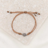 A taupe dainty string bracelet with a silver-tone Benedictine Medal and a charm, displayed on a textured background.