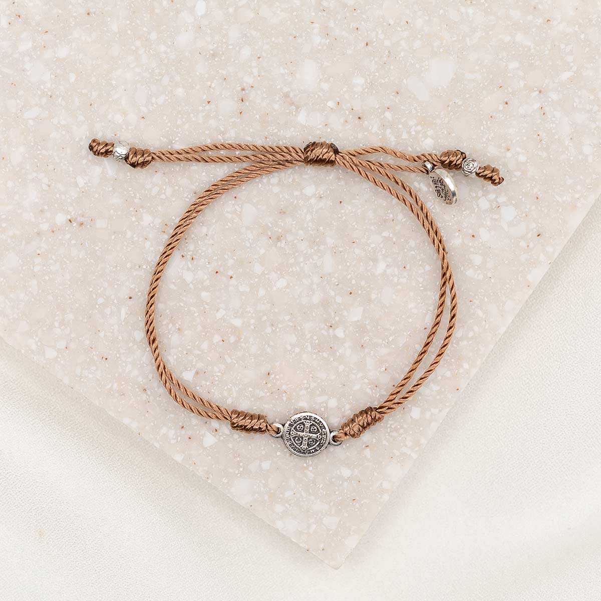 A taupe dainty string bracelet with a silver-tone Benedictine Medal and a charm, displayed on a textured background.