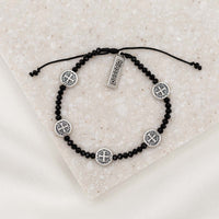 A black and silver Stellar Blessings Night Sky crystal blessing bracelet with adjustable cording, featuring silver-tone Benedictine medals and a 'blessed' charm tag, displayed on a textured background.