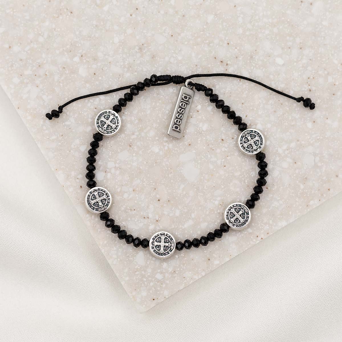 A black and silver Stellar Blessings Night Sky crystal blessing bracelet with adjustable cording, featuring silver-tone Benedictine medals and a 'blessed' charm tag, displayed on a textured background.