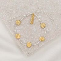 A clear crystal and gold blessing bracelet with adjustable cording, featuring gold-tone Benedictine medals and a 'blessed' charm tag, displayed on a textured background.