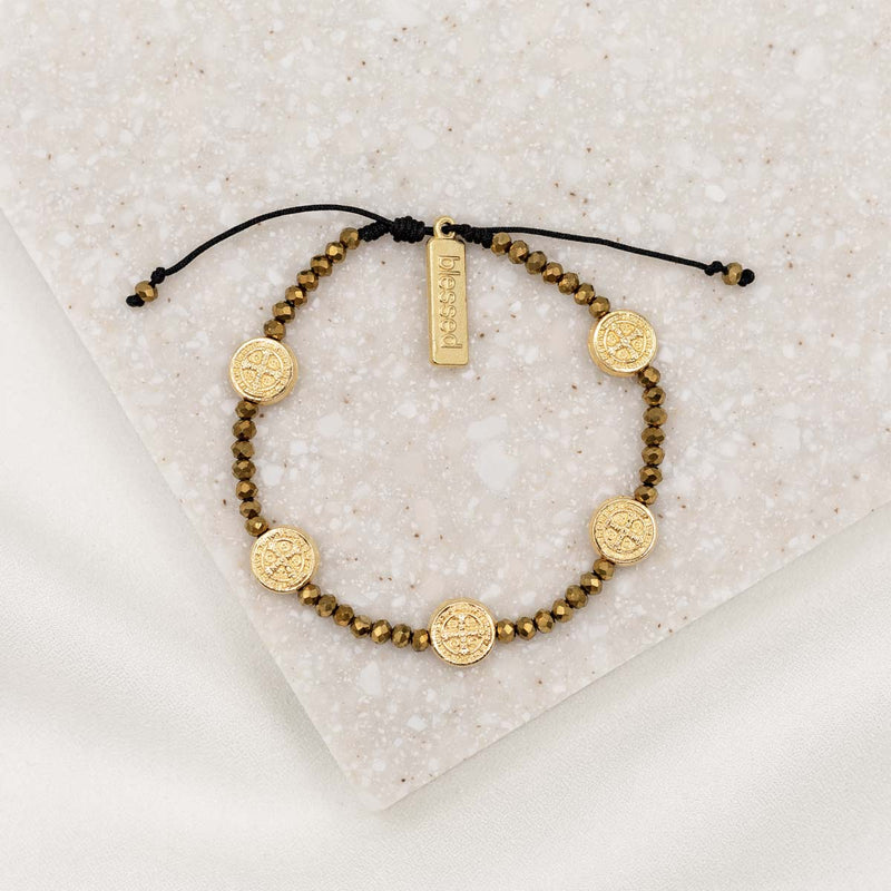 A black and gold gratitude crystal blessing bracelet with adjustable cording, featuring silver or gold-tone Benedictine medals and a 'blessed' charm tag, displayed on a textured background.
